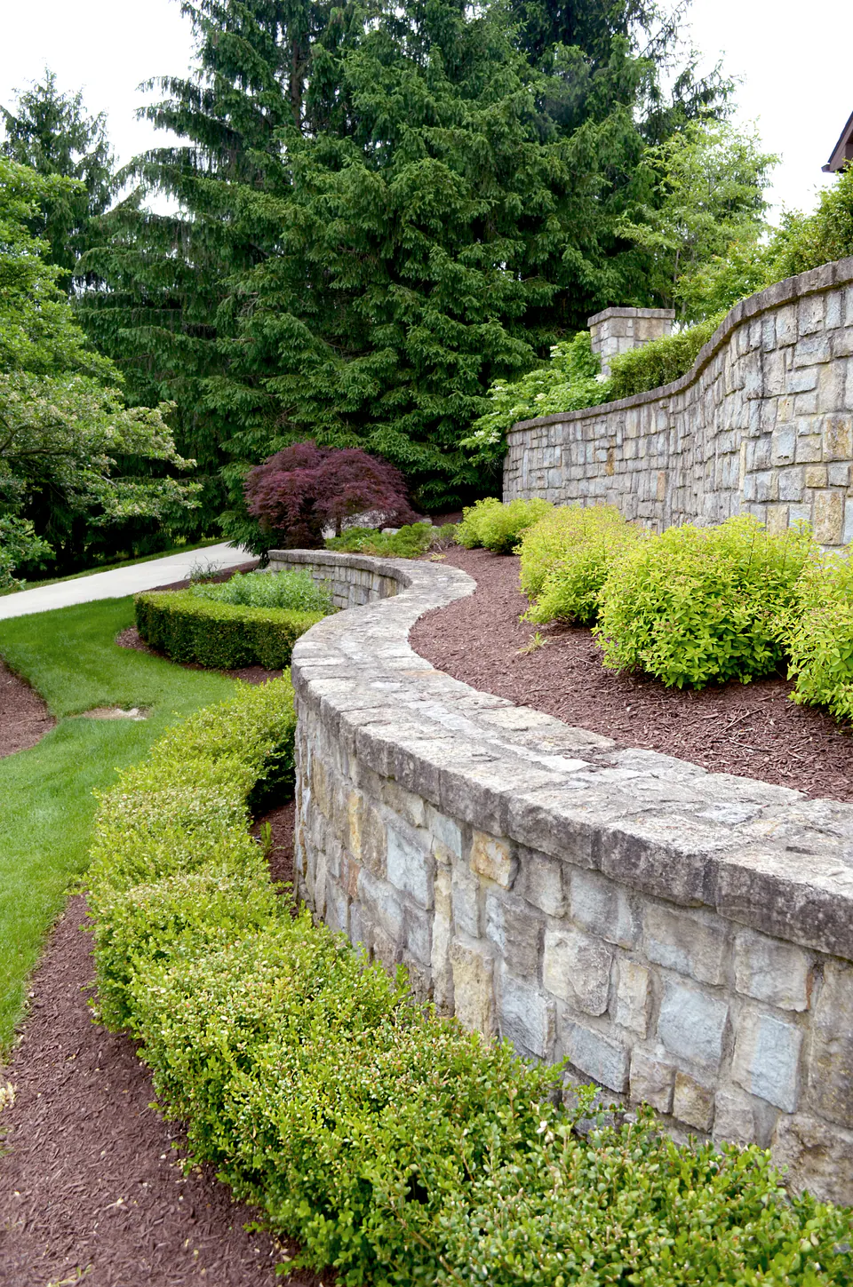 Long curved stone retaining wall with a stone cap above a clipped hedge and lawn